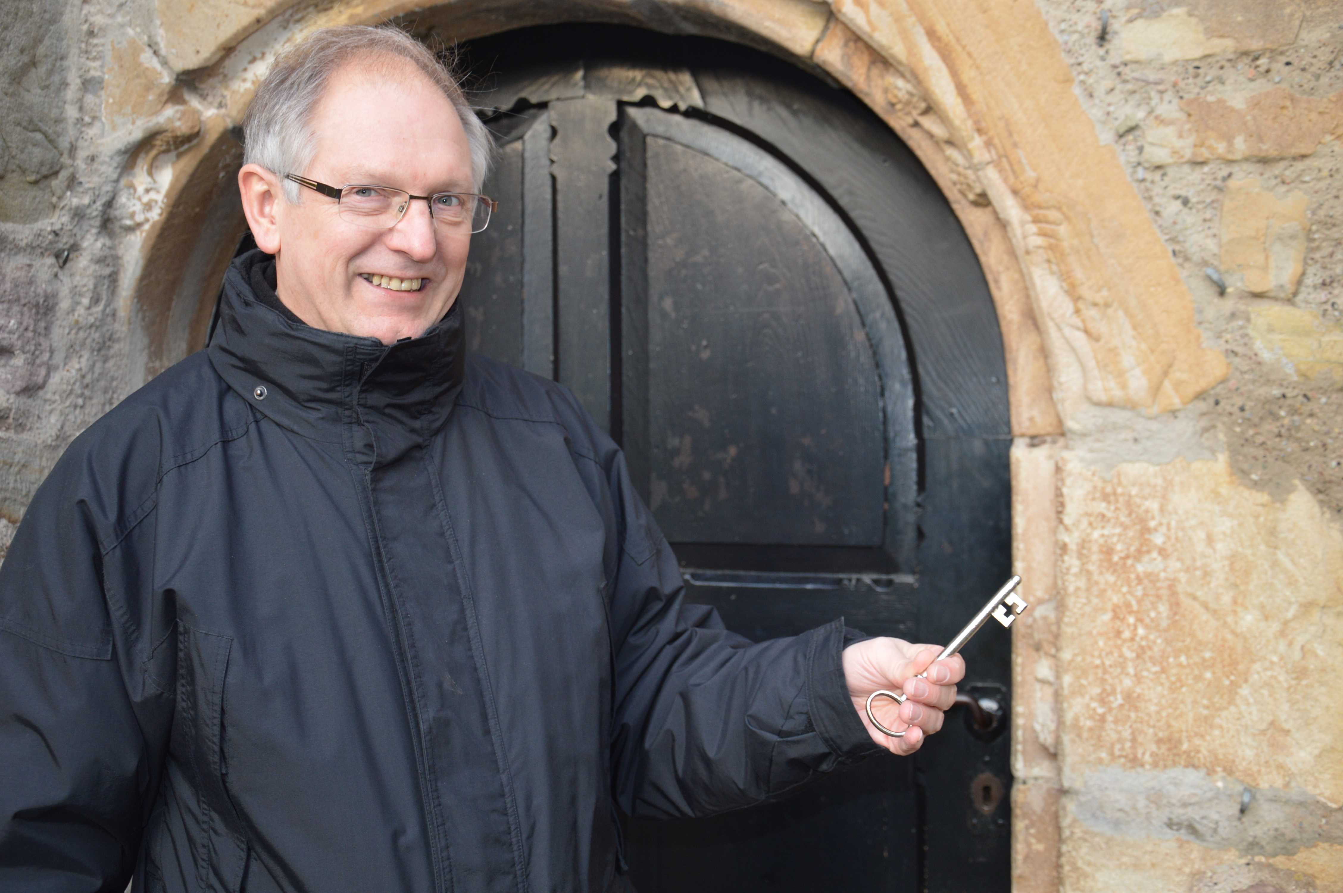 Pastor Reuning zeigt mit dem Kirchenschlüssel auf die in Sandstein eingearbeitete Hand, die einen Schlüssel hält.